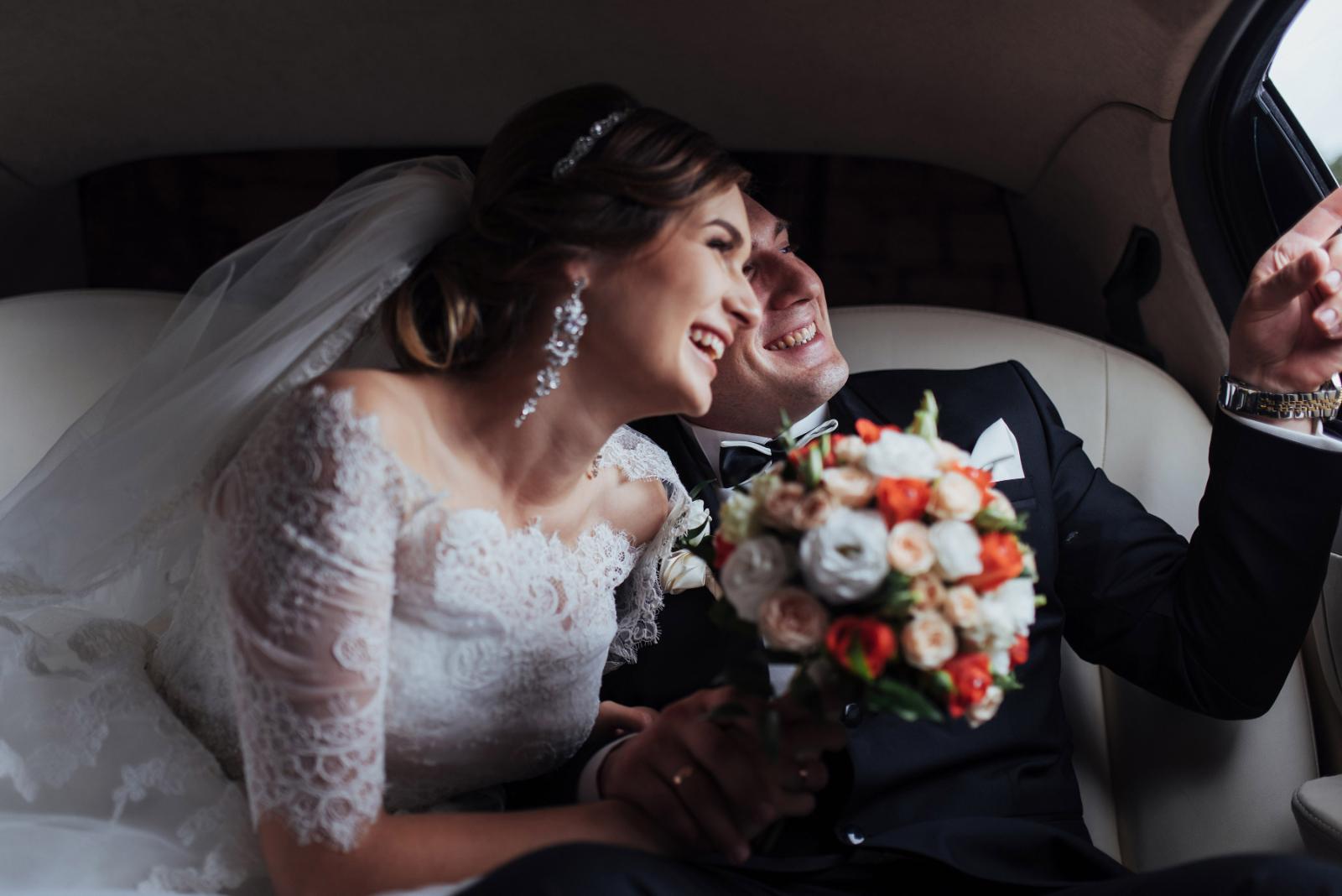 Glückliches Brautpaar lacht gemeinsam auf der Rückbank einer Hochzeitslimousine mit buntem Brautstrauß